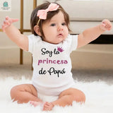 Baby wearing a white bodysuit with "Soy la princesa de Papá" text and a pink crown graphic, sitting on a soft rug with a pink bow headband.